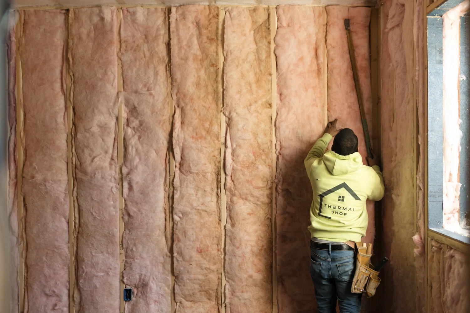 Installer in a Thermal Shop hoodie fitting pink fiberglass batts into the stud cavities of a framed wall