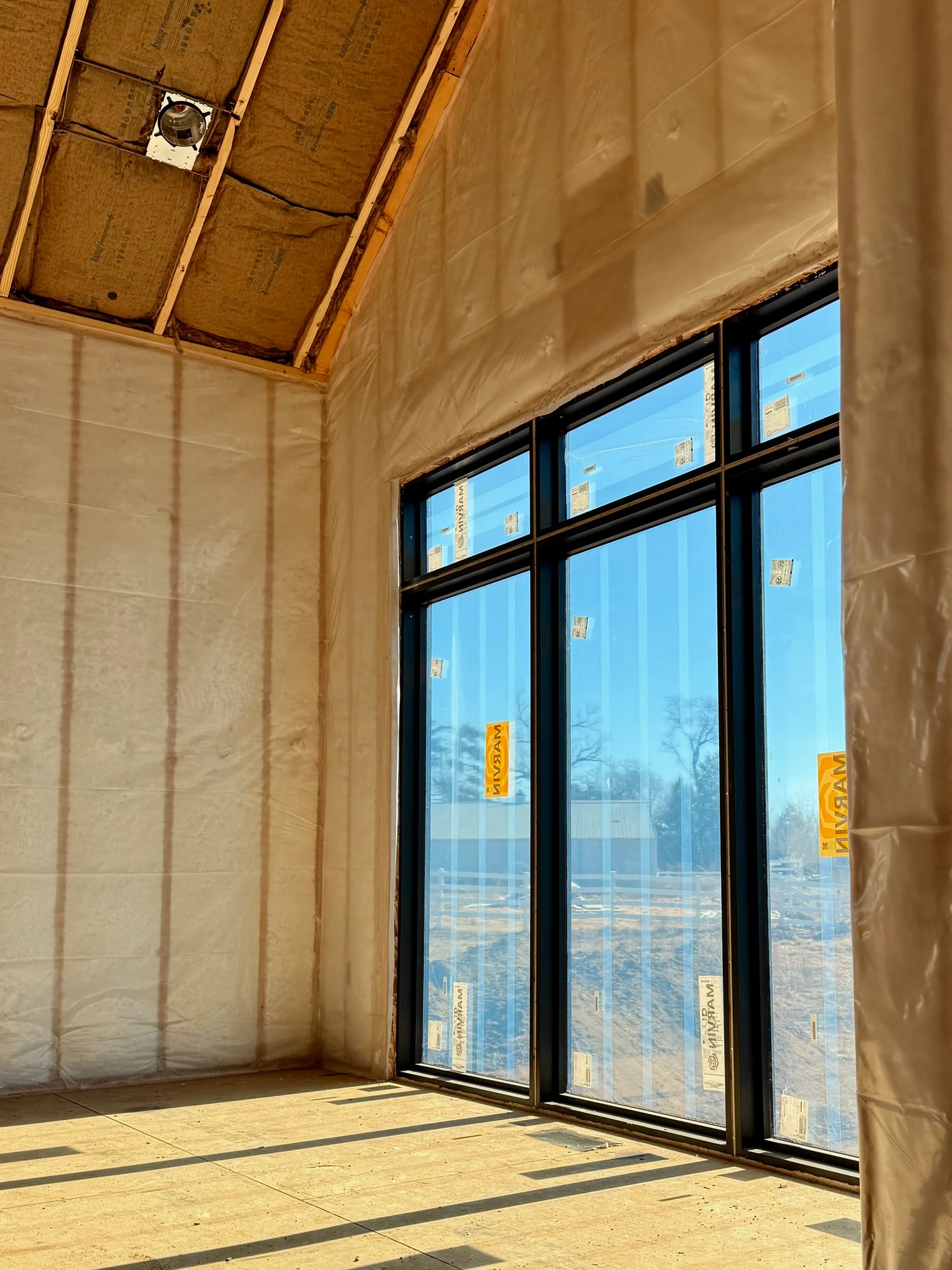 Room insulated behind netting, ready for drywall, with tall black-framed windows letting in low sun