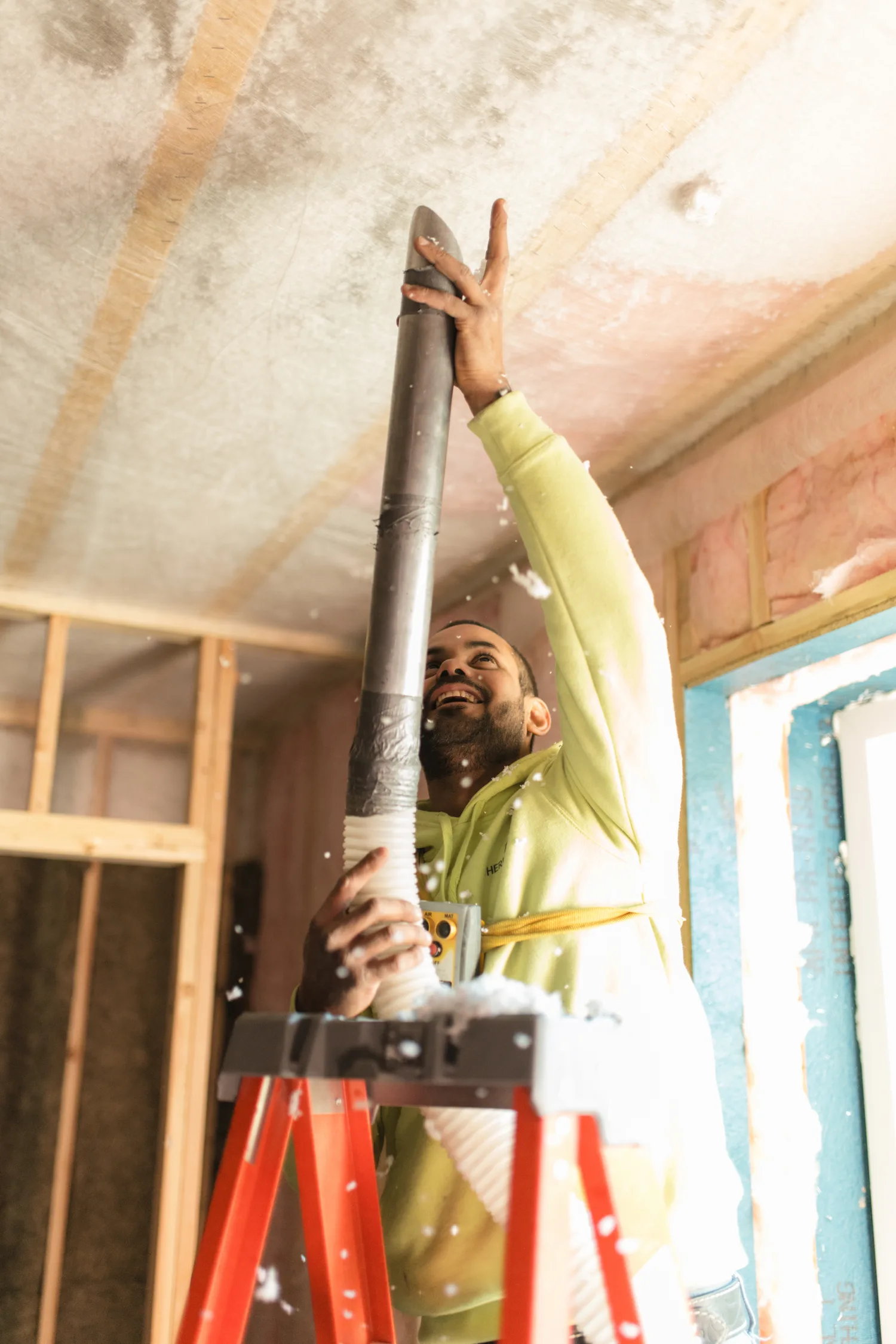 Smiling Thermal Shop installer on a red stepladder, holding the blowing hose up to a netted ceiling as loose insulation drifts down