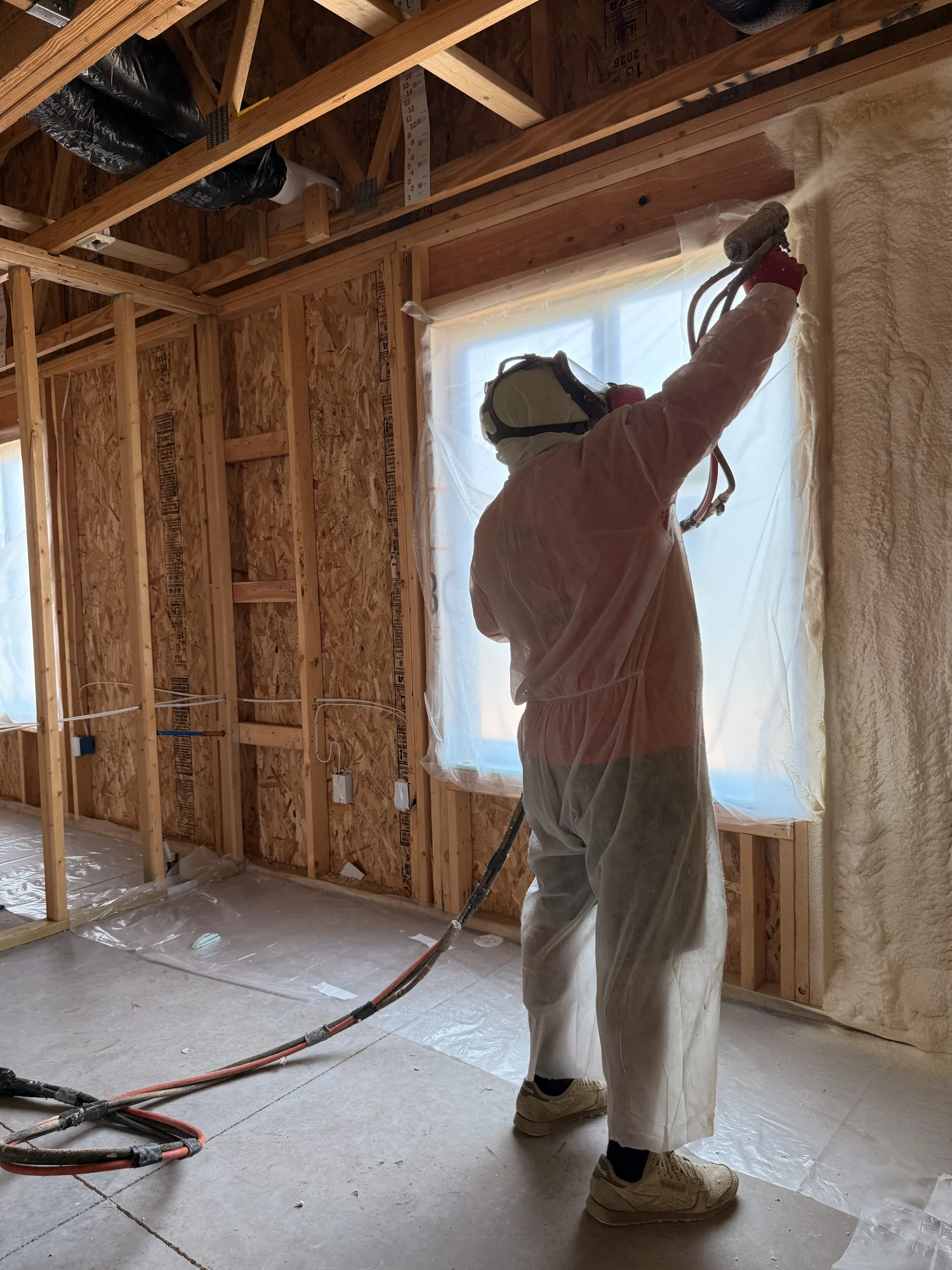 Installer in a protective suit spraying foam insulation around a window, silhouetted against the light